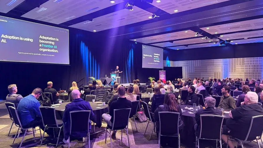 A Roadshow presenter on stage at a full convention hall, with slides reading Adoption is using AI, adaptation is becoming a Frontier AI organisation